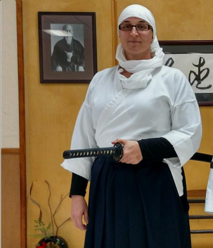 A woman stands wearing a white headscarf and martial arts robe, holding a sword