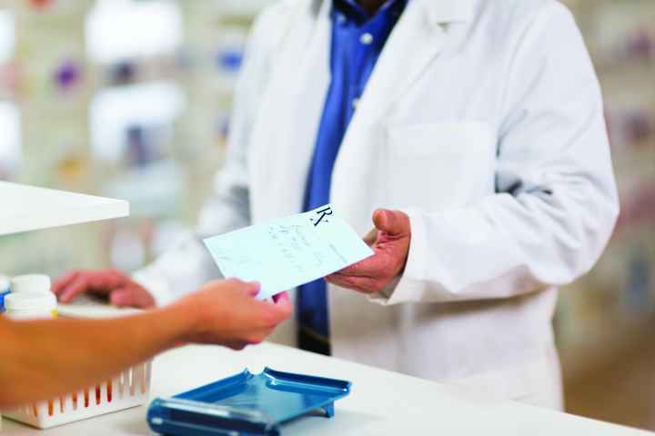 Photo: cropped photo of a pharmacist in a white coat behind a counter receiving a paper prescription from an outstretched hand 