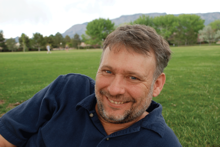 Photo: A man with short hair and groomed facial hair in a navy blue polo shirt reclining on a field of grass, smiling at the camera
