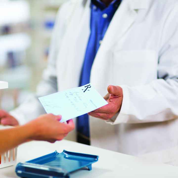 Photo: cropped photo of a pharmacist in a white coat behind a counter receiving a paper prescription from an outstretched hand 