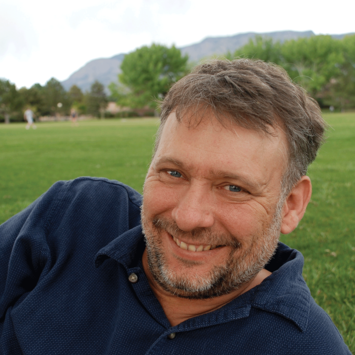 Photo: A man with short hair and groomed facial hair in a navy blue polo shirt reclining on a field of grass, smiling at the camera